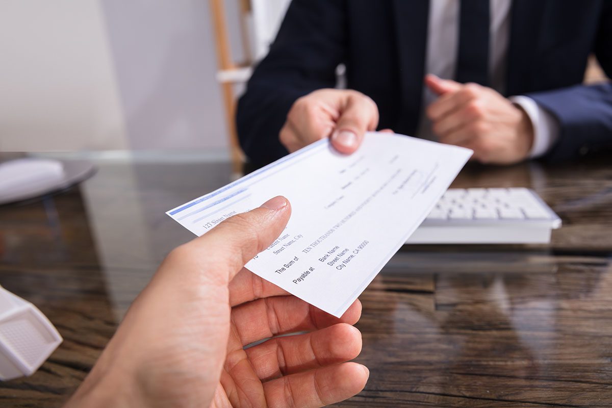 A person hands a check to another individual across a desk, with a keyboard and paperwork visible on the glass tabletop.
