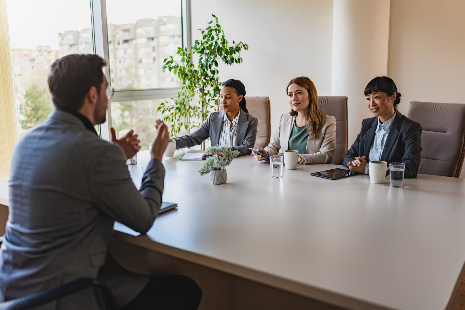 A man in a suit sits across from three women at a conference table, engaged in a discussion or interview in a modern office setting.