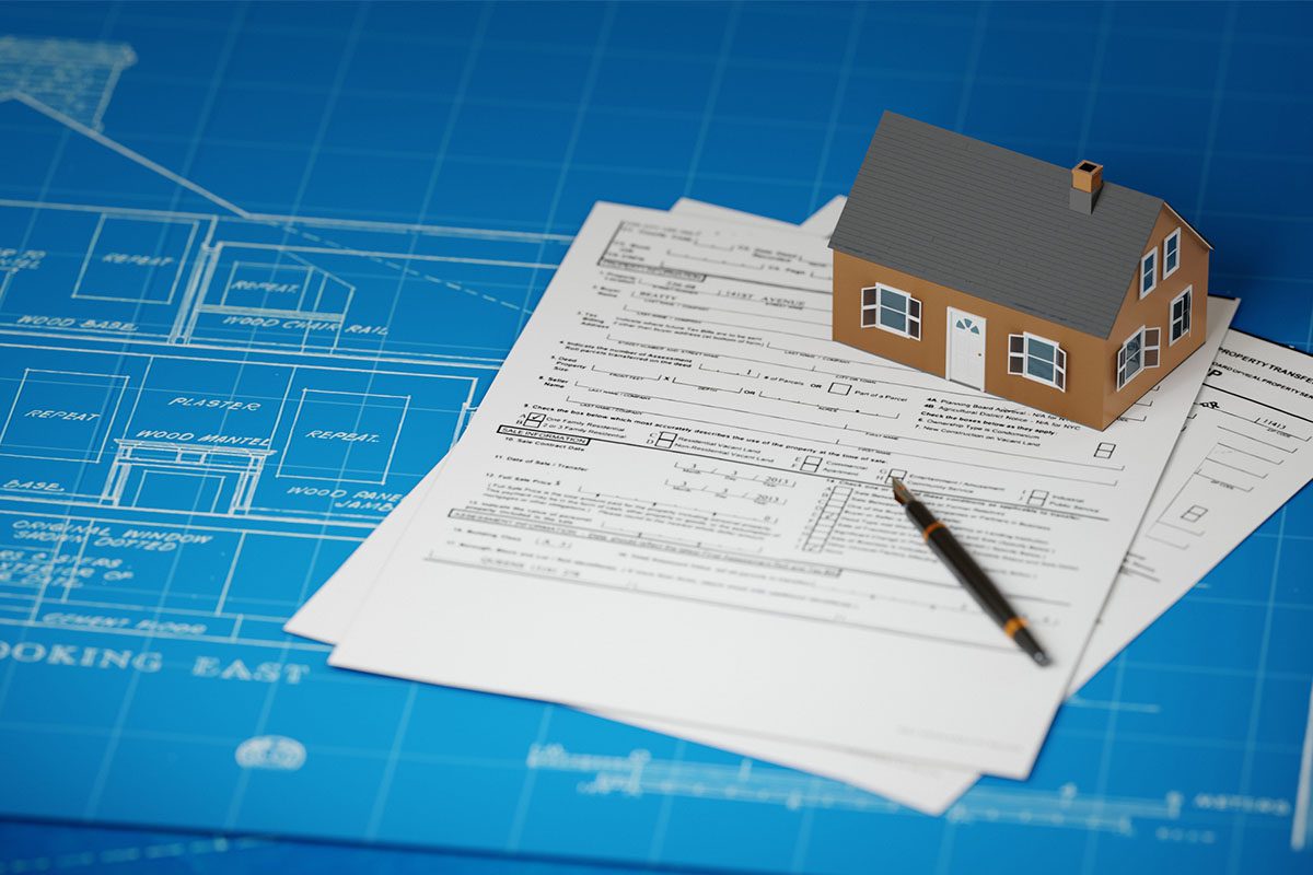 A small model house and a pen sit on top of several mortgage forms, placed on blue architectural plans.