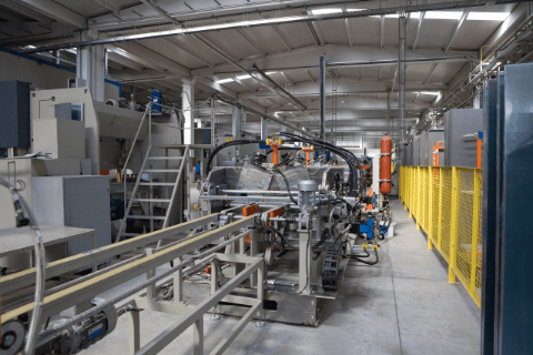 A modern industrial factory interior with automated machinery, conveyor belts, and metal structures under a high ceiling with fluorescent lighting.