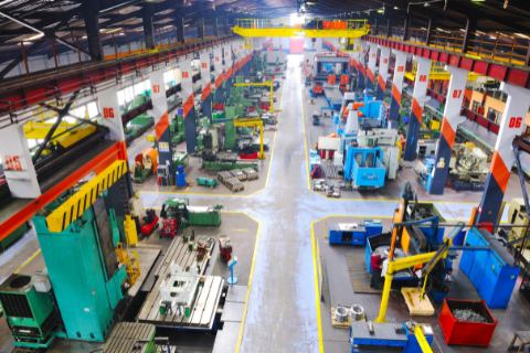 Large industrial factory interior with various heavy machinery, workstations, and equipment arranged in organized rows under a high ceiling.