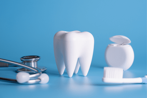 A model of a tooth stands on a blue surface next to a stethoscope, dental floss container, and toothbrush.