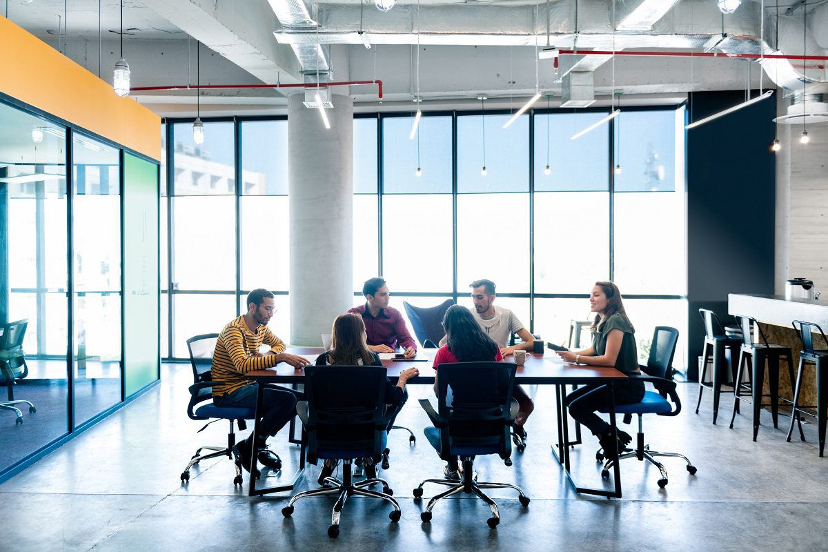 Six people sit around a table in a modern office meeting room, engaged in discussion, with large windows and abundant natural light in the background.