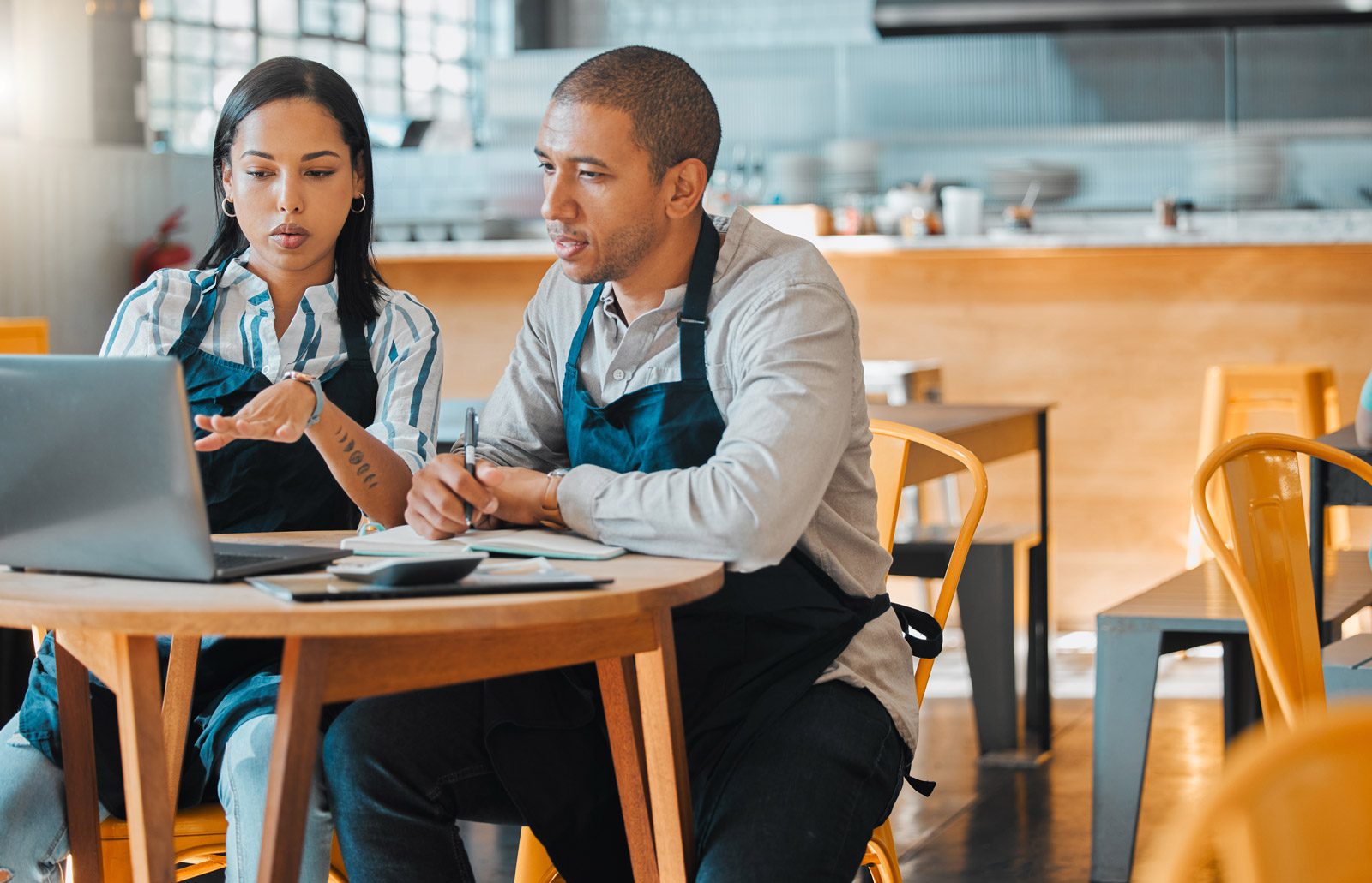 Two restaurant workers wearing aprons sit at a table, looking at a laptop and discussing something, with notepads and cups on the table in a modern dining area.