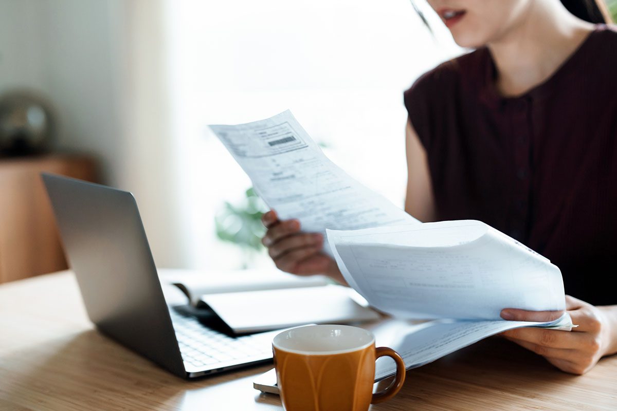 A person sits at a desk with a laptop, reviewing and holding several sheets of paper, with a coffee mug nearby.