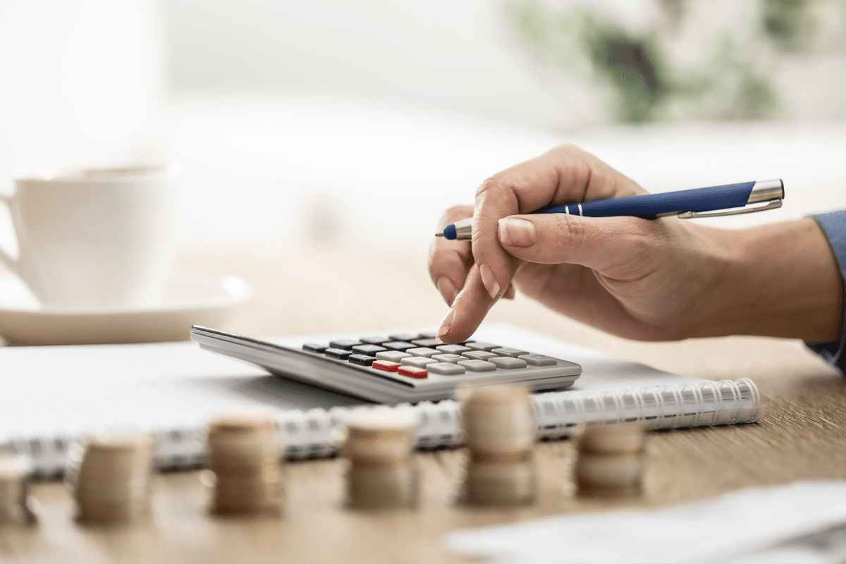 A person uses a calculator on a desk with coins stacked in the foreground and a notebook and coffee cup nearby.