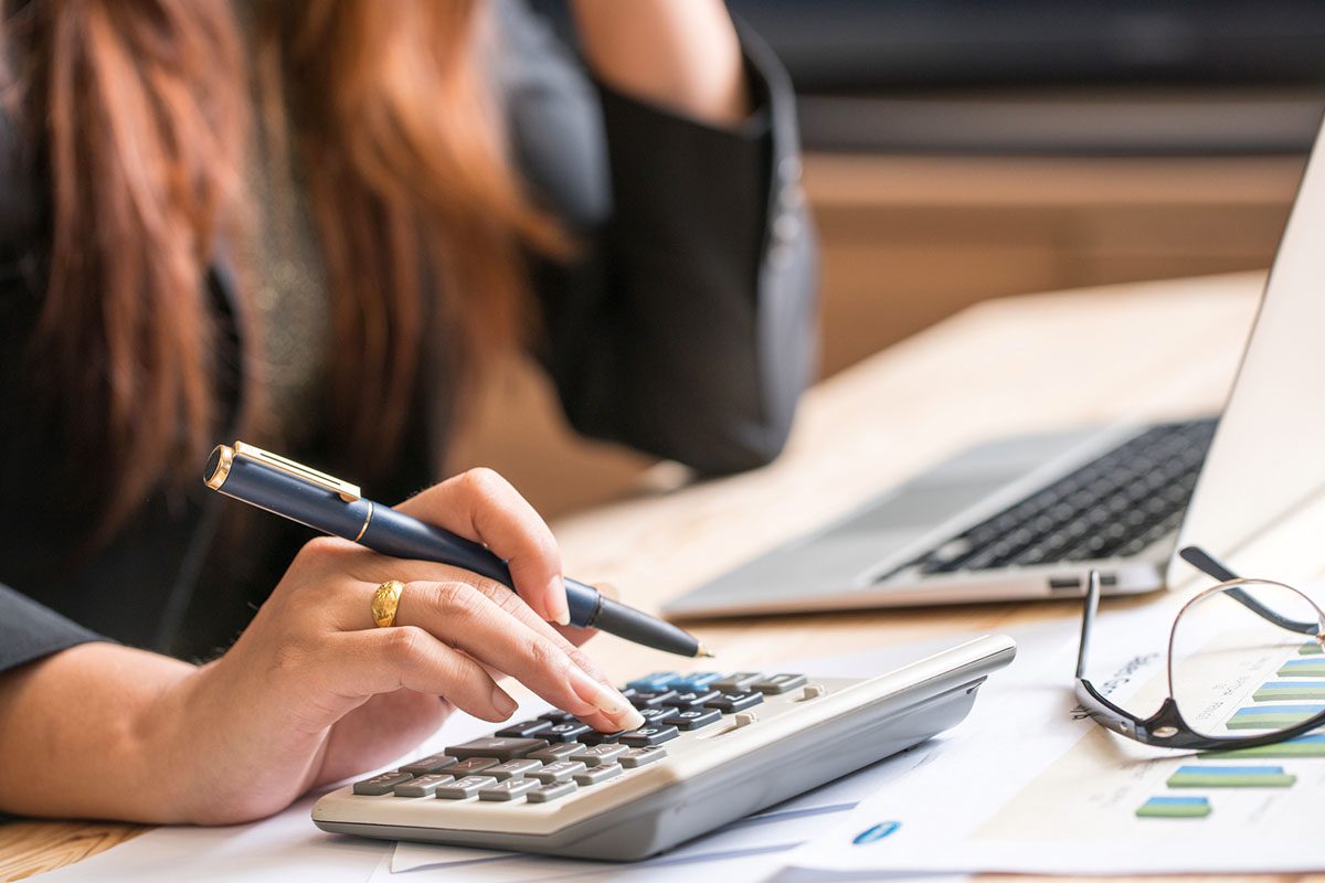 A person uses a calculator and holds a pen at a desk with papers, a laptop, and eyeglasses.