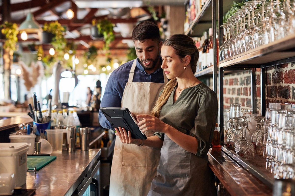 Two restaurant workers stand behind the bar, looking at a tablet together, surrounded by glasses and bottles on shelves in a well-lit setting.