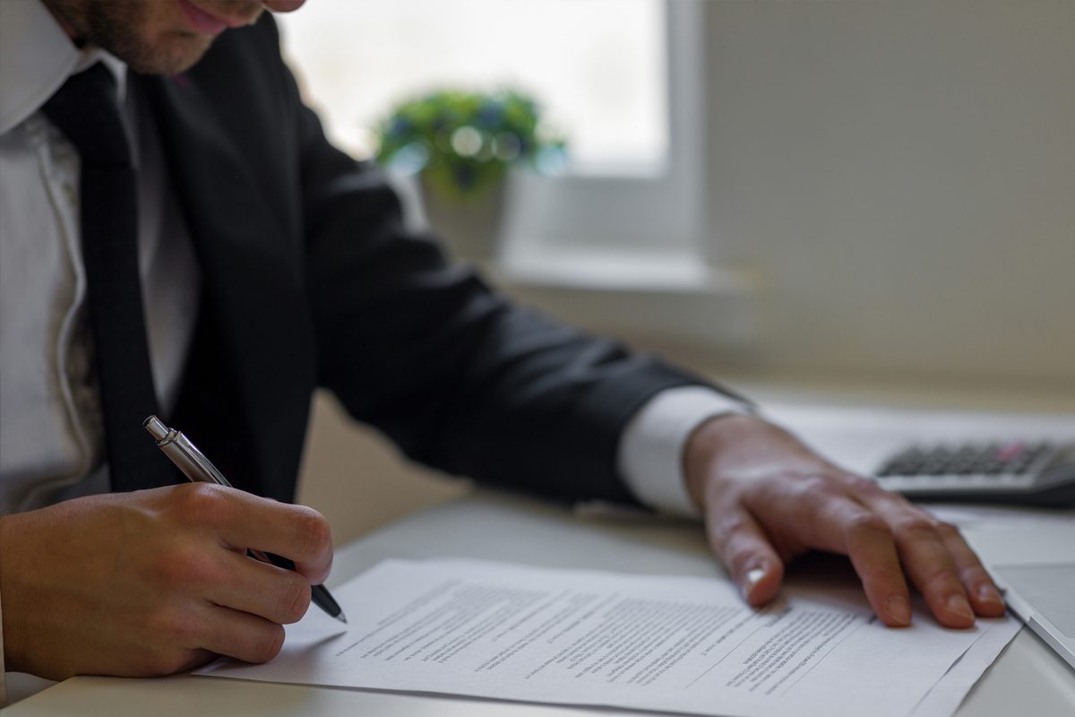 A person in a suit sits at a desk, holding a pen and signing a document, with a calculator and a small plant in the background.