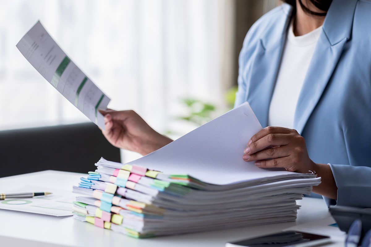 A person in a light blue blazer sorts through a large stack of documents with colorful paper clips at a desk.