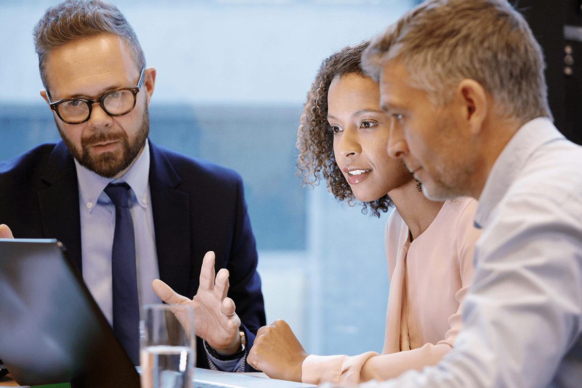 Three professionals sit at a table, looking at a laptop screen, engaged in a business discussion.
