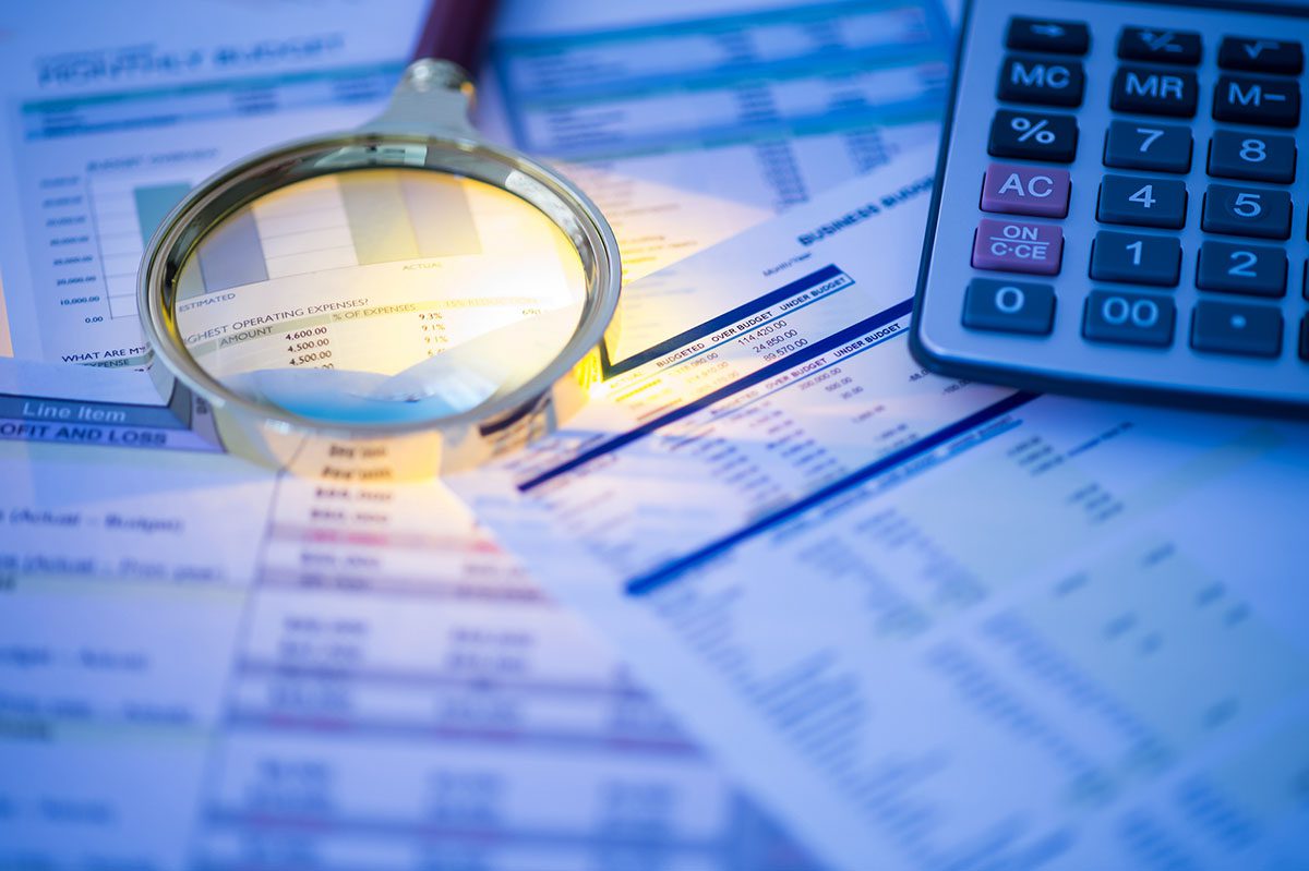A magnifying glass, financial documents, and a calculator are arranged on a table, with the magnifying glass highlighting part of a spreadsheet.