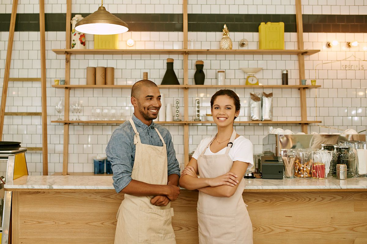 Two baristas wearing aprons stand behind a wooden counter in a modern coffee shop with tiled walls and shelves displaying various items.