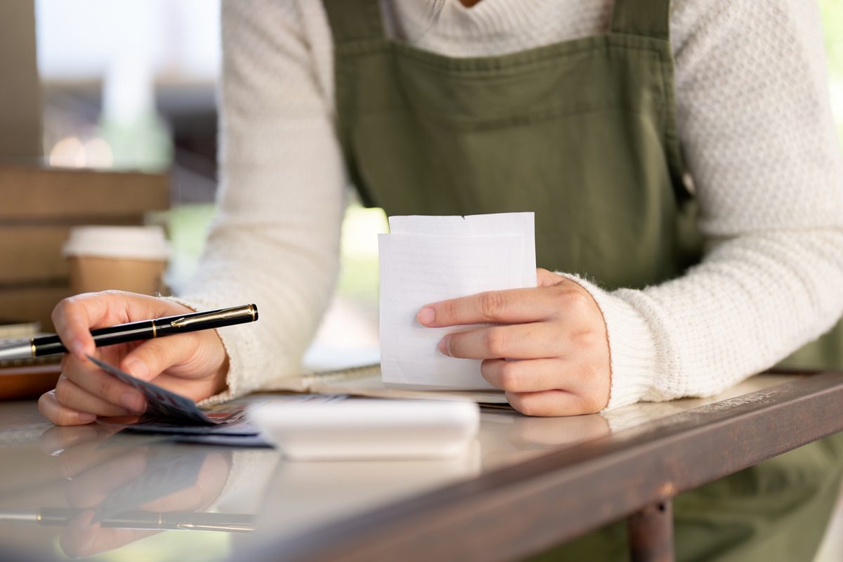 A person in a green apron reviews a receipt and holds a pen at a table with a credit card, a notebook, and a coffee cup in the background.