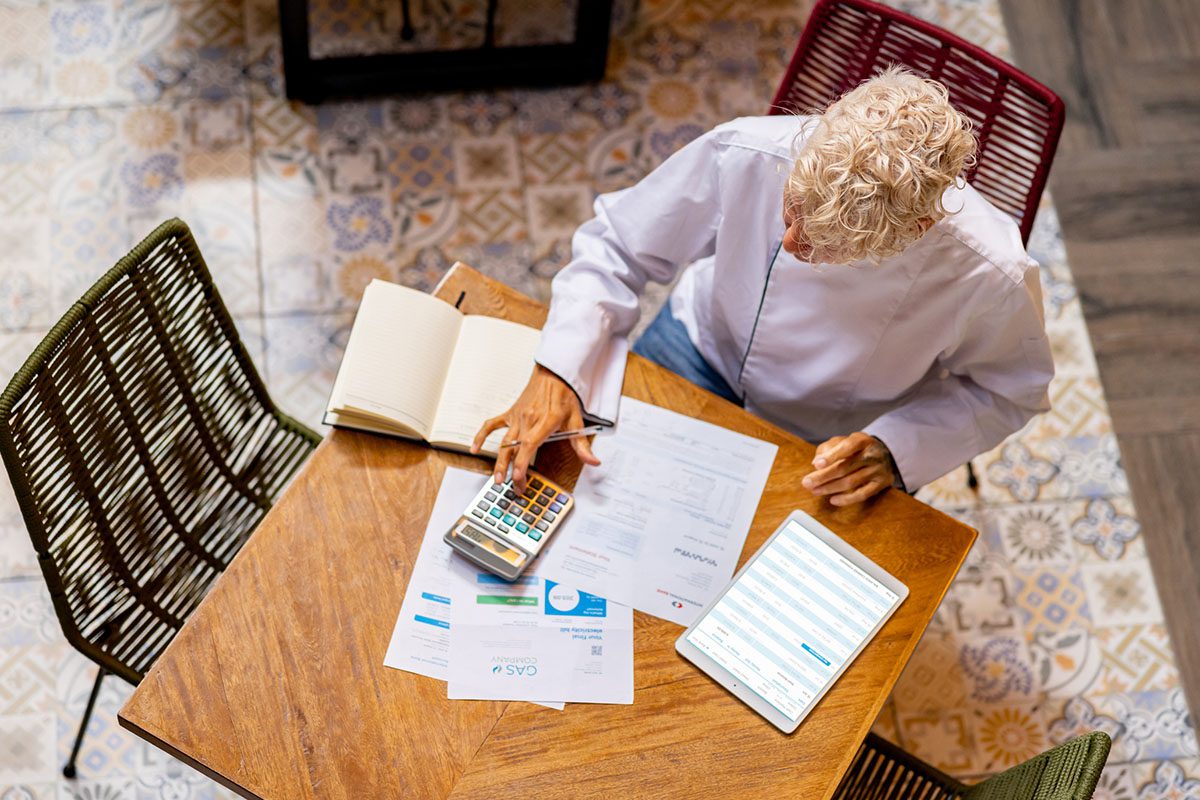 An older adult with gray hair sits at a table with papers, a notebook, a calculator, and a tablet, appearing to review financial documents.