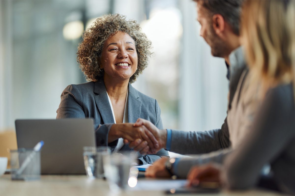 A woman in a business suit smiles and shakes hands with a man across a table, with a laptop and documents visible in an office setting.