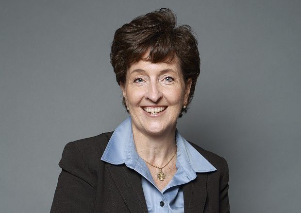 A middle-aged woman with short brown hair, wearing a dark blazer over a light blue collared shirt, smiles at the camera against a plain gray background.