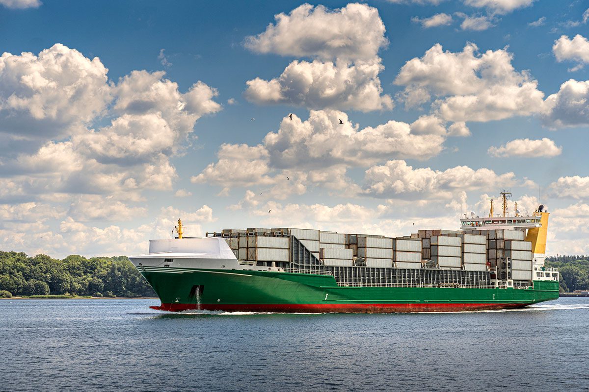 A large green and white cargo ship carrying stacked containers sails on a body of water under a partly cloudy sky.