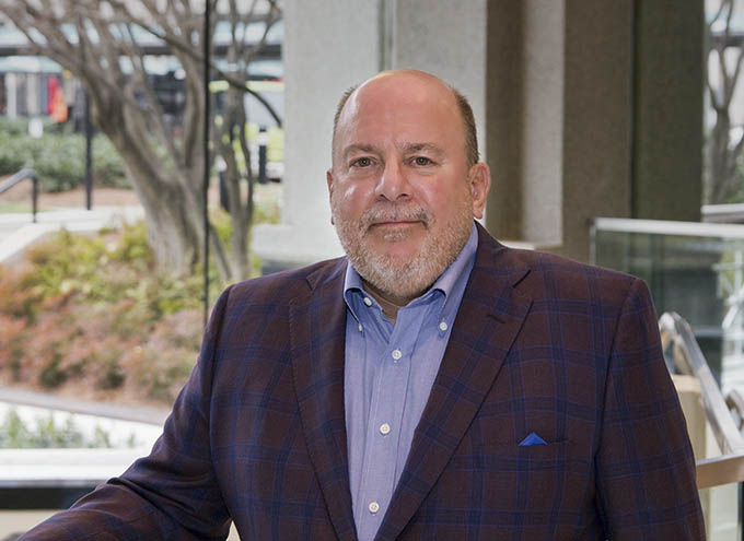 A middle-aged man with a beard and mustache, wearing a blue shirt and plaid blazer, stands indoors near a large window with outdoor greenery visible in the background.