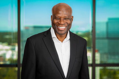 A man in a black suit jacket and white shirt stands smiling in front of large windows with a cityscape in the background.