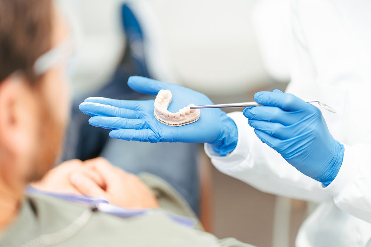 A dental professional wearing blue gloves holds a dental model with tweezers and shows it to a seated patient.