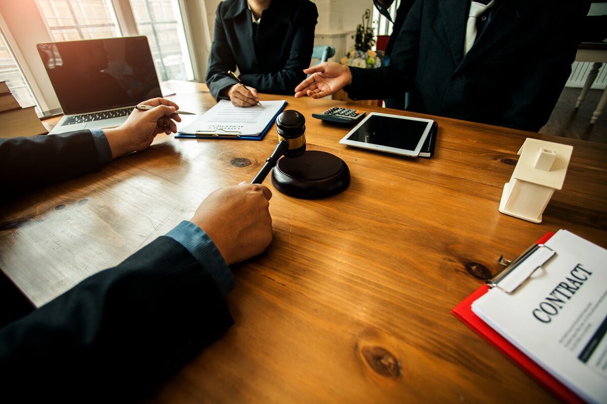 Three people in business attire sit around a wooden table with a gavel, contract papers, a tablet, a laptop, and a model house, suggesting a legal or business meeting.