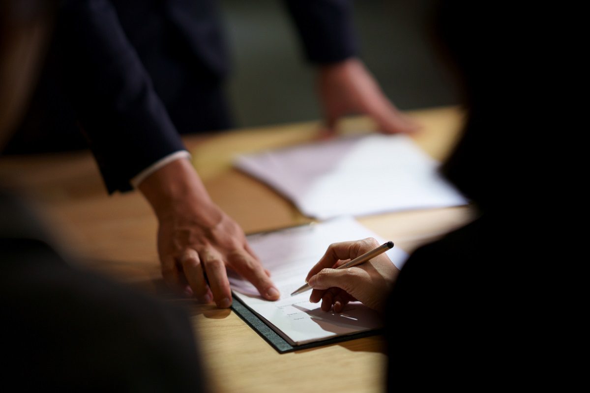 A person signs a document on a clipboard while another individual points to the paperwork on a table.