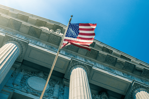 Upward view of the United States flag flying in front of a classical building with tall columns under a clear blue sky.