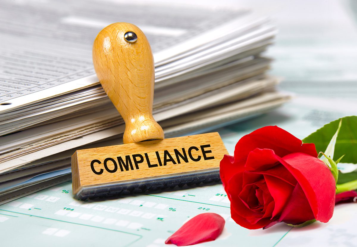A rubber stamp labeled COMPLIANCE sits on paperwork beside a red rose with scattered petals.