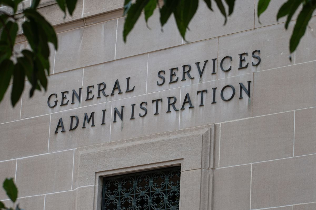 The words General Services Administration are displayed on the exterior wall of a stone building, partially framed by green leaves.