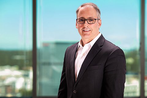 A man in a dark suit and glasses stands in front of a large window overlooking a cityscape on a sunny day.