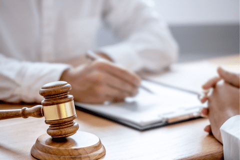 A wooden gavel rests on a desk while two people have a discussion, with one person writing on a clipboard.