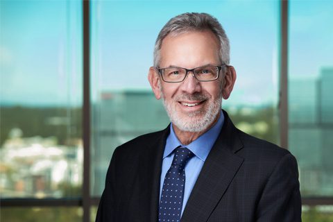 An older man with gray hair, a beard, and glasses is wearing a dark suit, blue shirt, and polka dot tie, standing in front of large office windows with a city view.