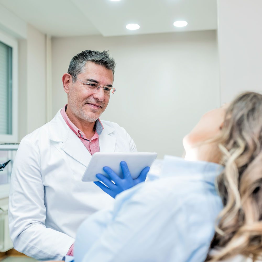 A male doctor in a white coat holds a tablet and talks to a patient lying back in a medical examination chair.