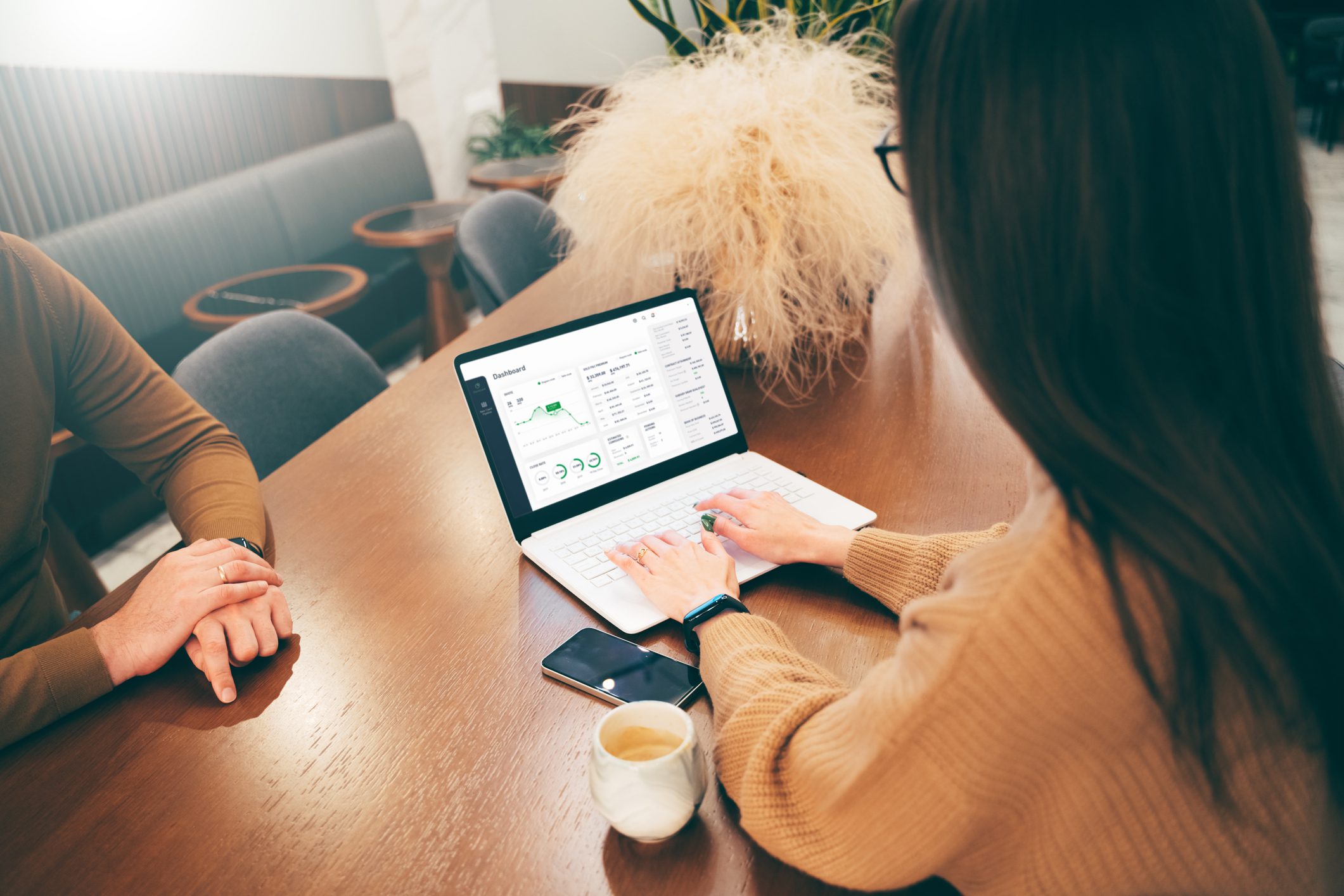 A woman reviews charts and data on a laptop at a wooden table with a coffee cup, phone, and another person seated nearby.