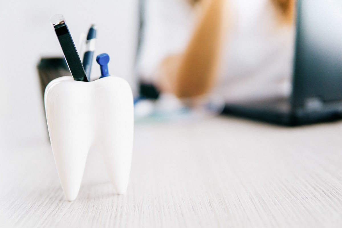 A tooth-shaped pen holder containing pens and a pushpin is on a desk, with a blurred person and a laptop in the background.