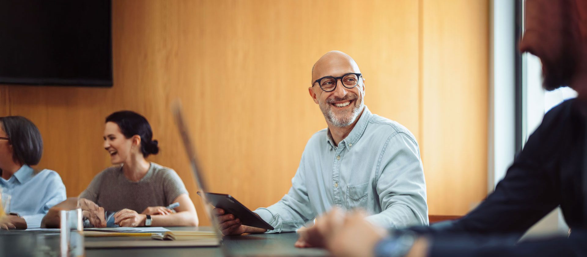 Sitting at a conference room table, a bald man with glasses laughs at someone obscured in the foreground