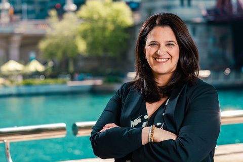 A woman with long dark hair, wearing a black blazer, stands outdoors with arms crossed, smiling in front of a body of water and trees.