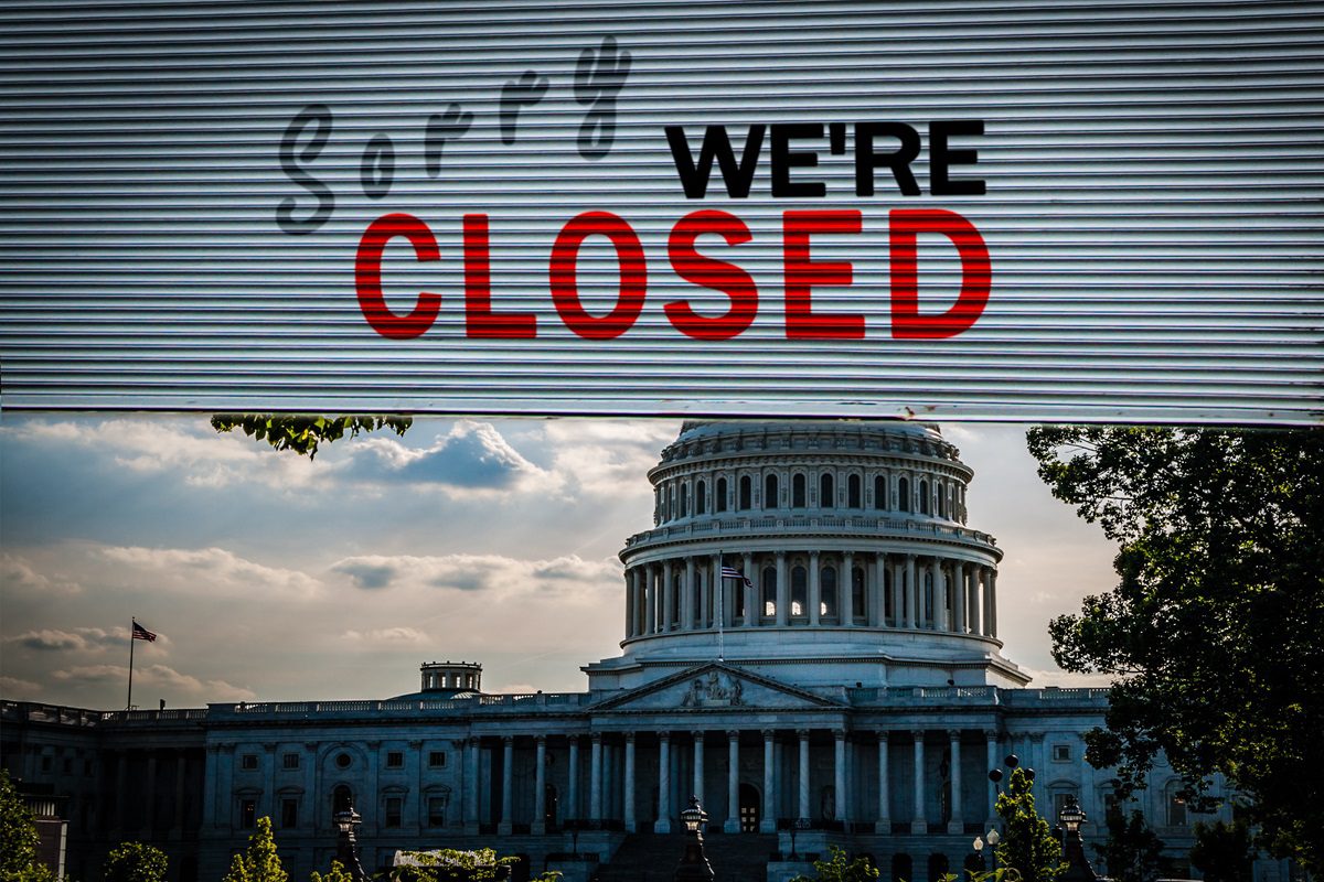 A metal shutter with Sorry Were Closed partially covers a view of the U.S. Capitol building under a cloudy sky.