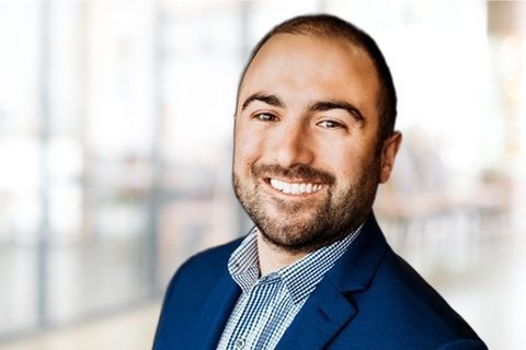 A man with short brown hair and a beard, wearing a blue blazer and checked shirt, smiles at the camera in a bright, blurred office setting.