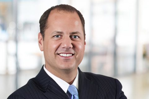 A man in a dark suit and tie is smiling, standing in a bright, blurred office setting.