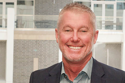 A middle-aged man with short gray hair and a trimmed beard smiles at the camera, wearing a blue blazer and a checked shirt, with a modern building in the background.