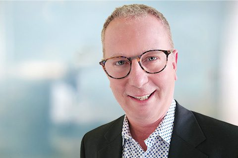 A middle-aged man with short, light hair, wearing glasses, a dark blazer, and a white patterned shirt, smiling in front of a blurred background.