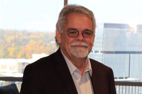 An older man with white hair, a beard, and glasses is wearing a dark suit and standing indoors with city buildings and trees visible through the window behind him.