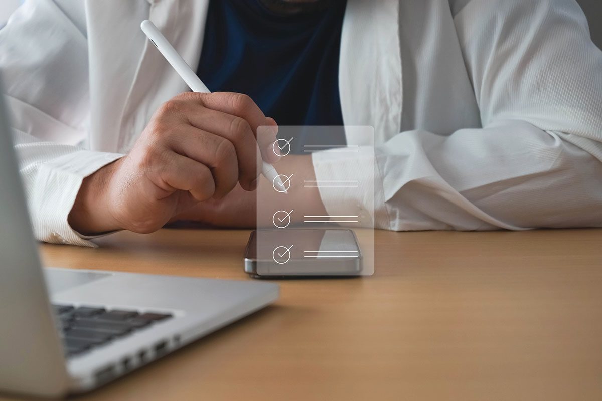 Person using a stylus on a smartphone with a digital checklist overlay, sitting at a desk with a laptop in the foreground.