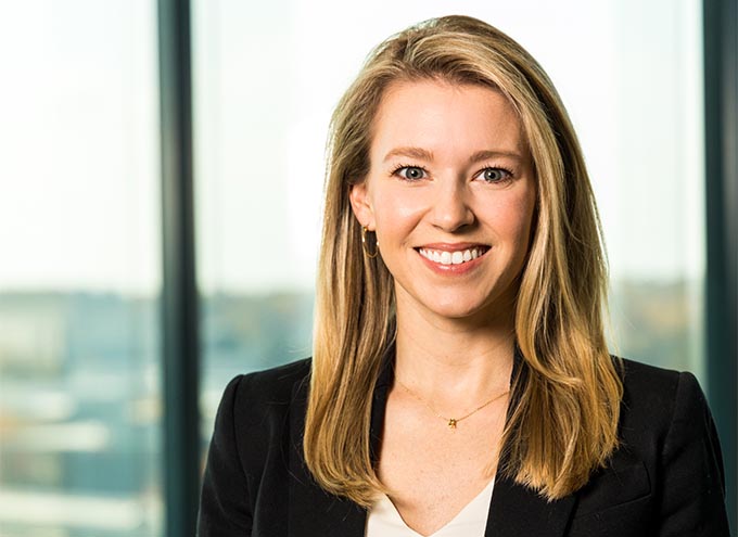 A woman with long blonde hair, wearing a black blazer and white top, smiles at the camera in front of large office windows.