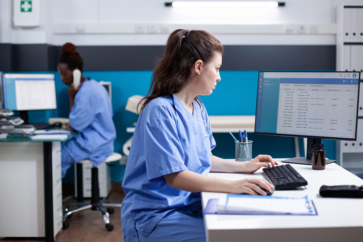 Two healthcare professionals in scrubs work in a medical office; one types at a computer while the other speaks on the phone in the background.