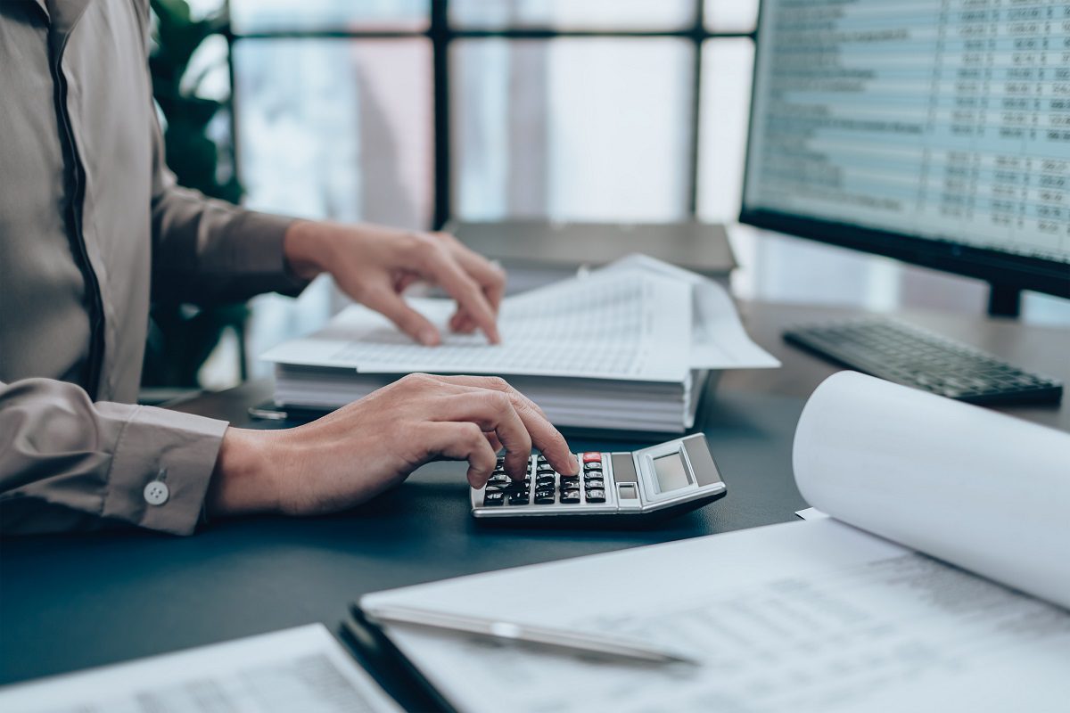 Person using a calculator and reviewing documents at a desk with financial spreadsheets on a computer monitor in an office setting.
