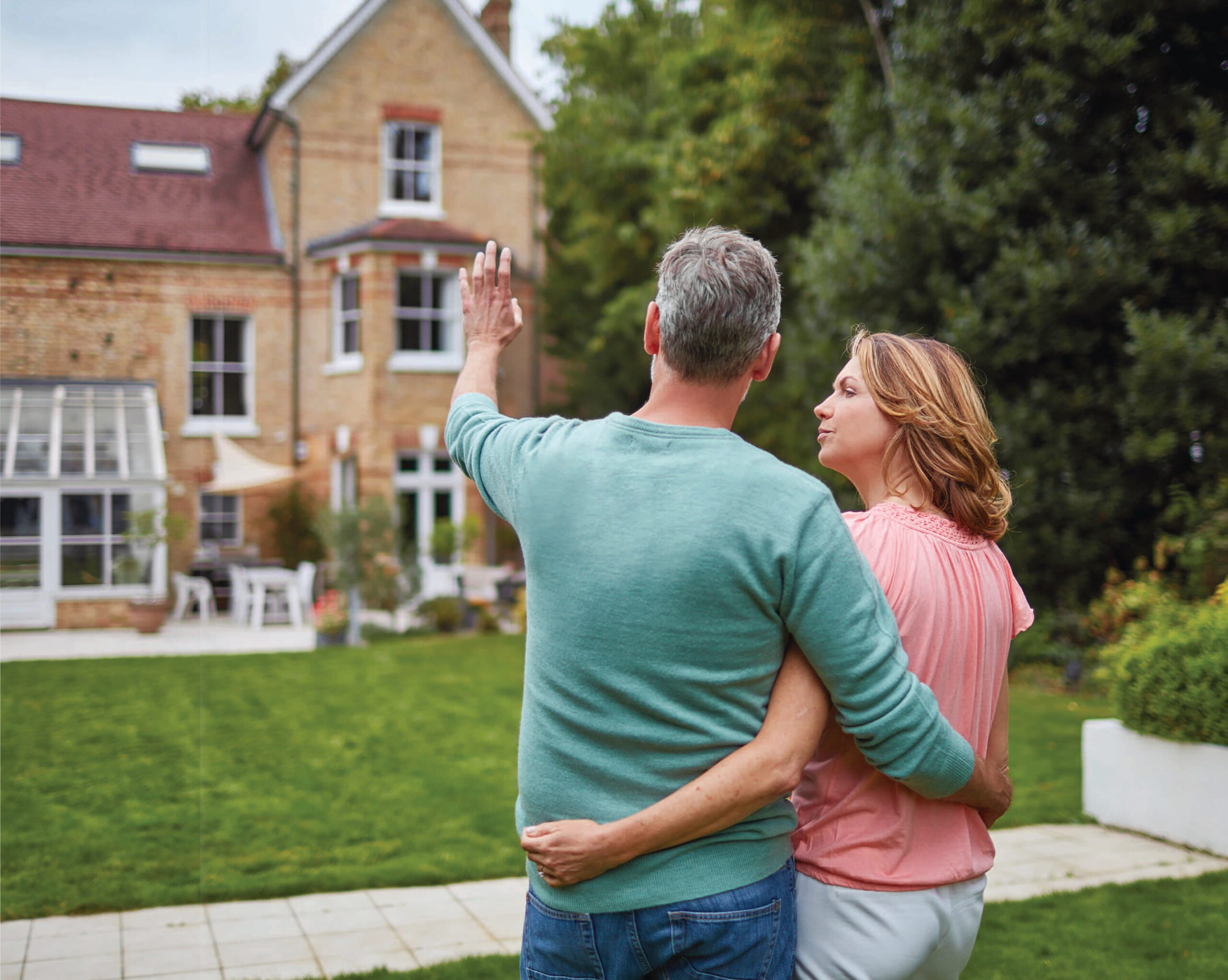 A man and woman stand arm in arm in a garden, facing and waving toward a large brick house.