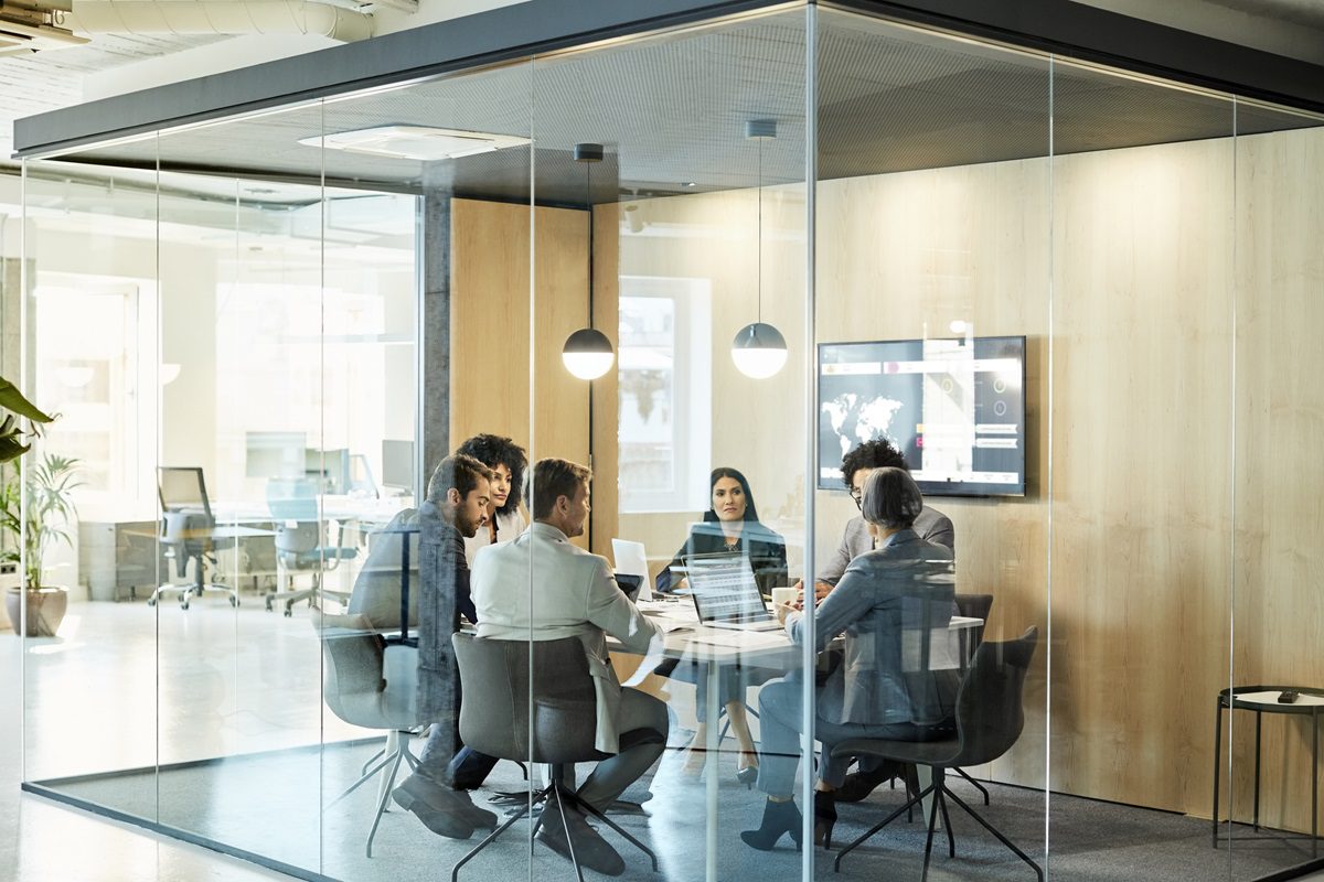 Six people sit around a table in a modern glass-walled conference room, engaged in a meeting. A large monitor displays charts on the wall.
